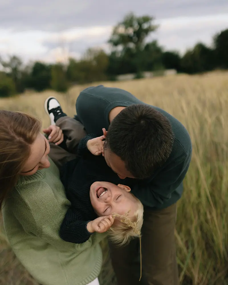 Föräldrar som busar och skrattar med sitt barn i högt gräs.